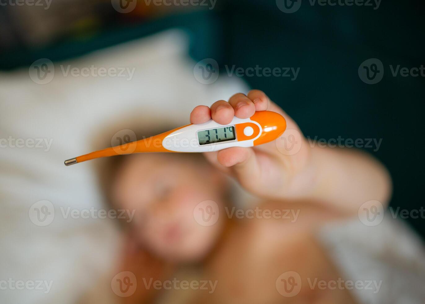 Child holding a digital thermometer. Blurred background with sick boy lying in bed. photo
