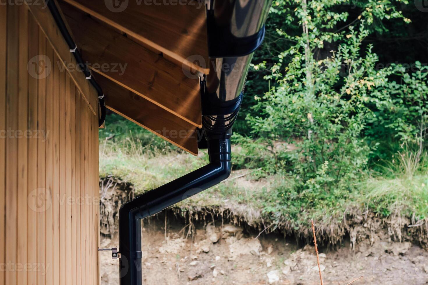Black drainage pipe under wooden roof near green vegetation in a natural landscape photo