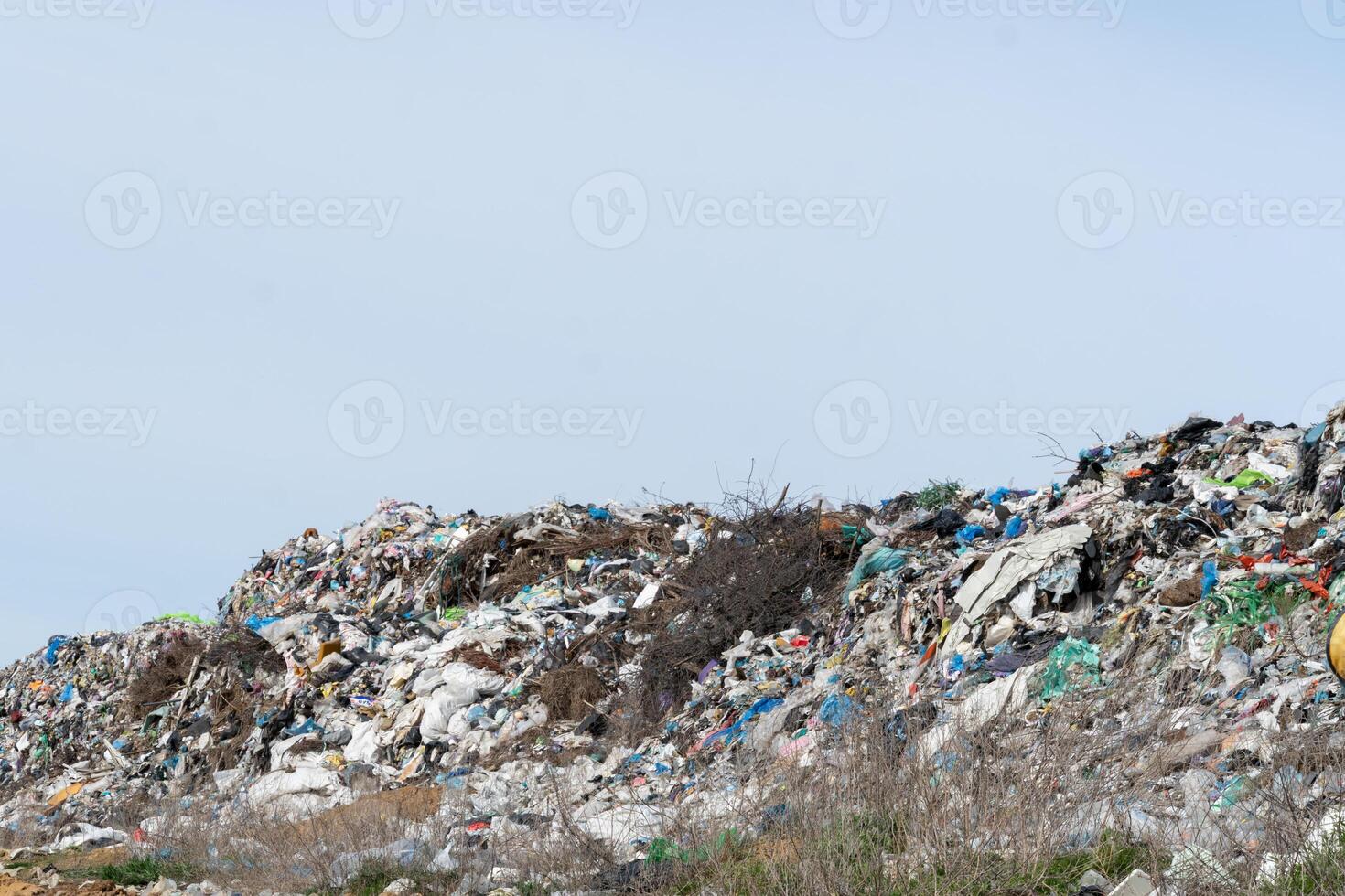 A large landfill area is filled with heaps of plastic waste and debris scattered across the landscape, illuminated by bright sunlight amidst a clear blue sky photo