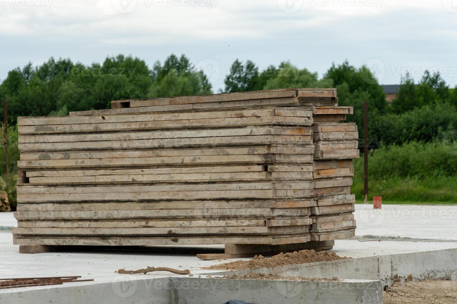A stack of wooden planks is neatly arranged on a concrete surface at a construction site surrounded by lush greenery under a cloudy sky photo