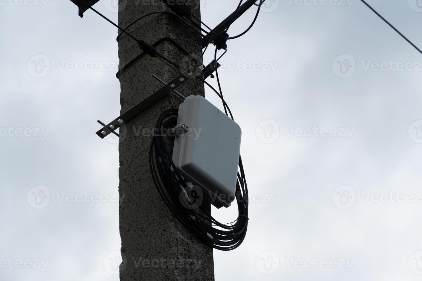Communication hardware is mounted on a utility pole with wires hanging down. The backdrop features a gray, cloud-filled sky indicative of overcast weather photo