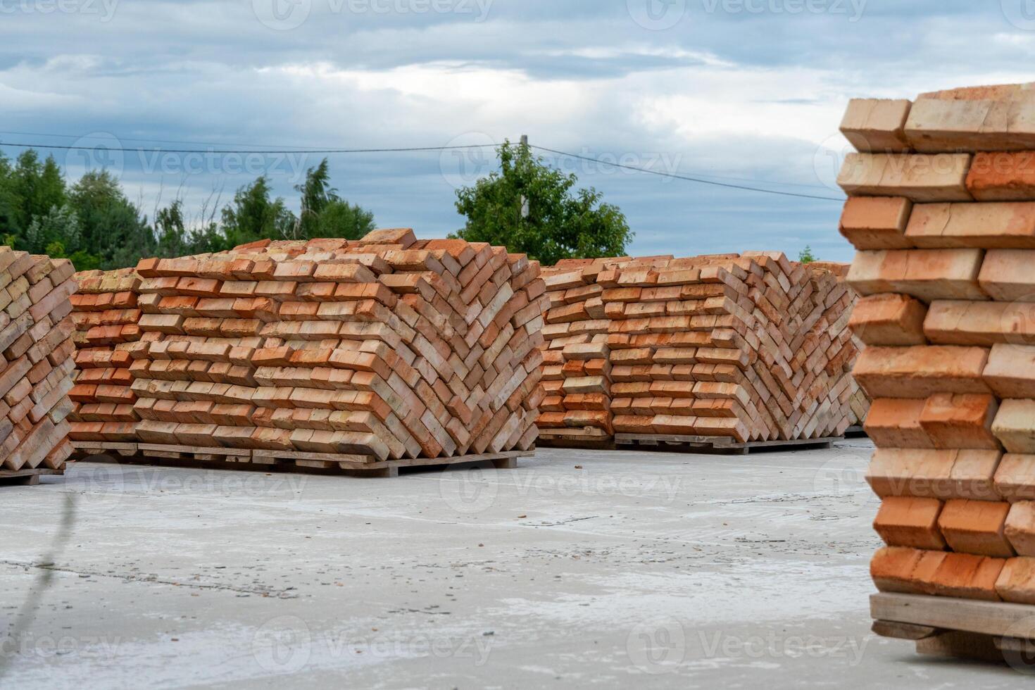 Piles of neatly arranged red bricks sit in a construction yard, ready for use. The sky is overcast, hinting at possible rain. Green trees provide a backdrop photo