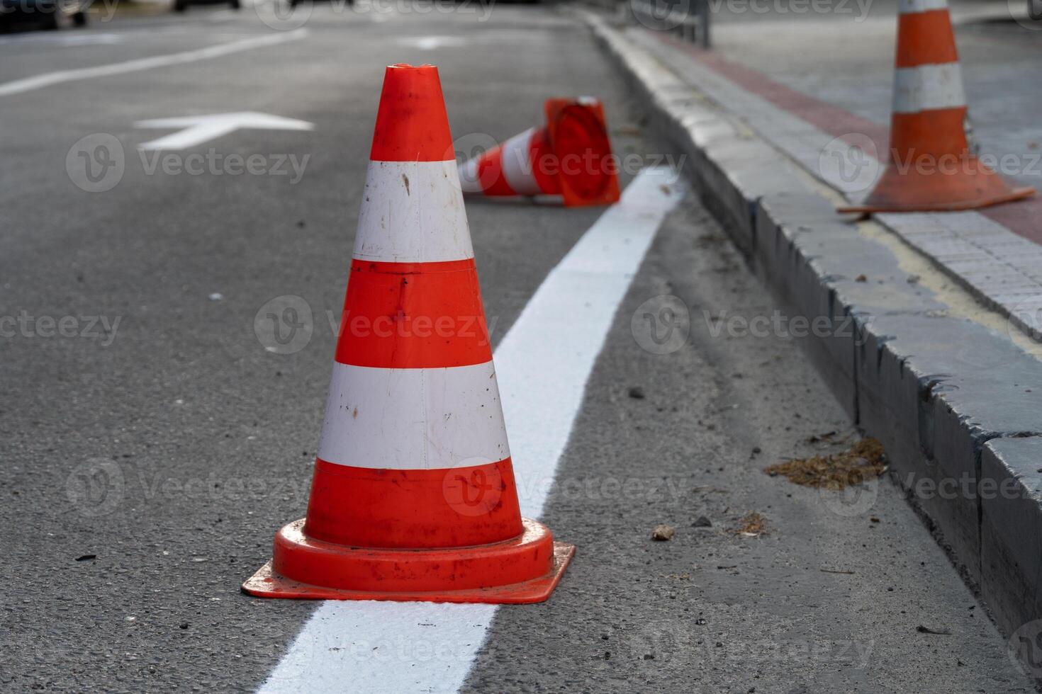 Bright orange and white traffic cones are placed along a freshly painted lane on a busy city street. Some cones are tipped over, indicating ongoing roadwork photo