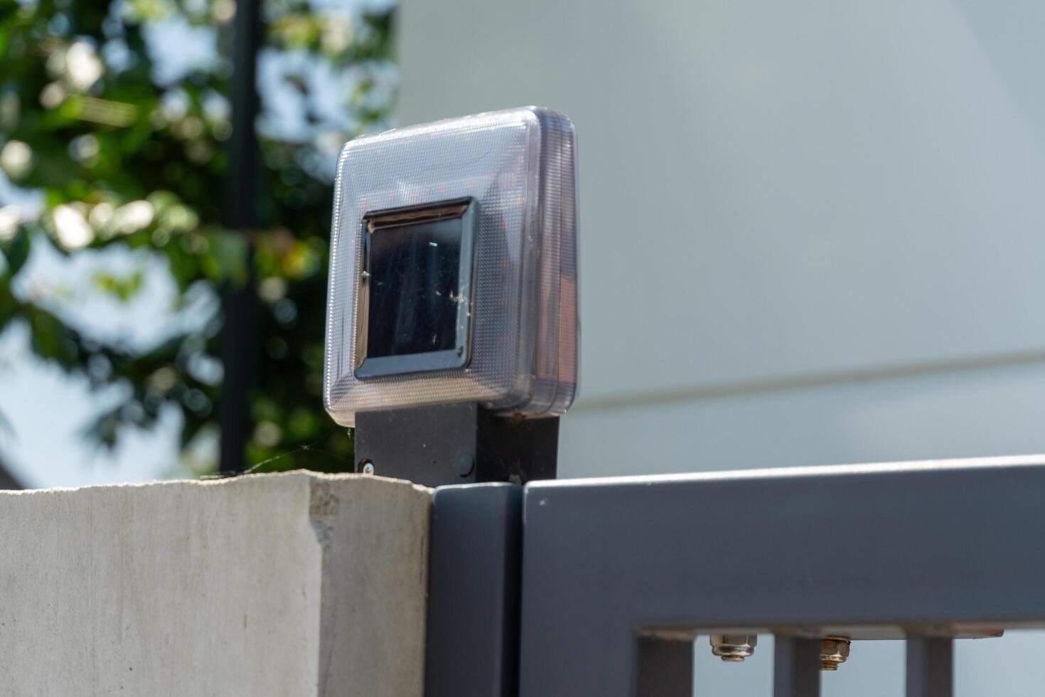A solar-powered light mounted on a gray metal gate provides illumination and security. It is situated in a sunny outdoor area with greenery in the background photo