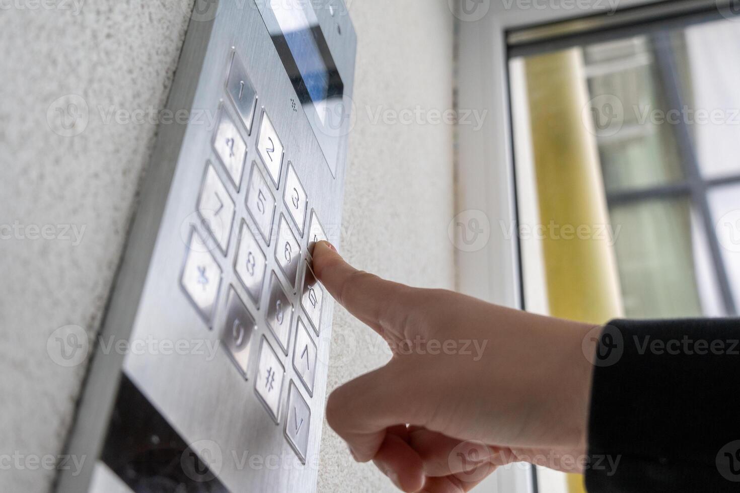 A person is pressing a button on a sleek security entry system attached to a wall in an urban setting while sunlight filters through nearby windows photo