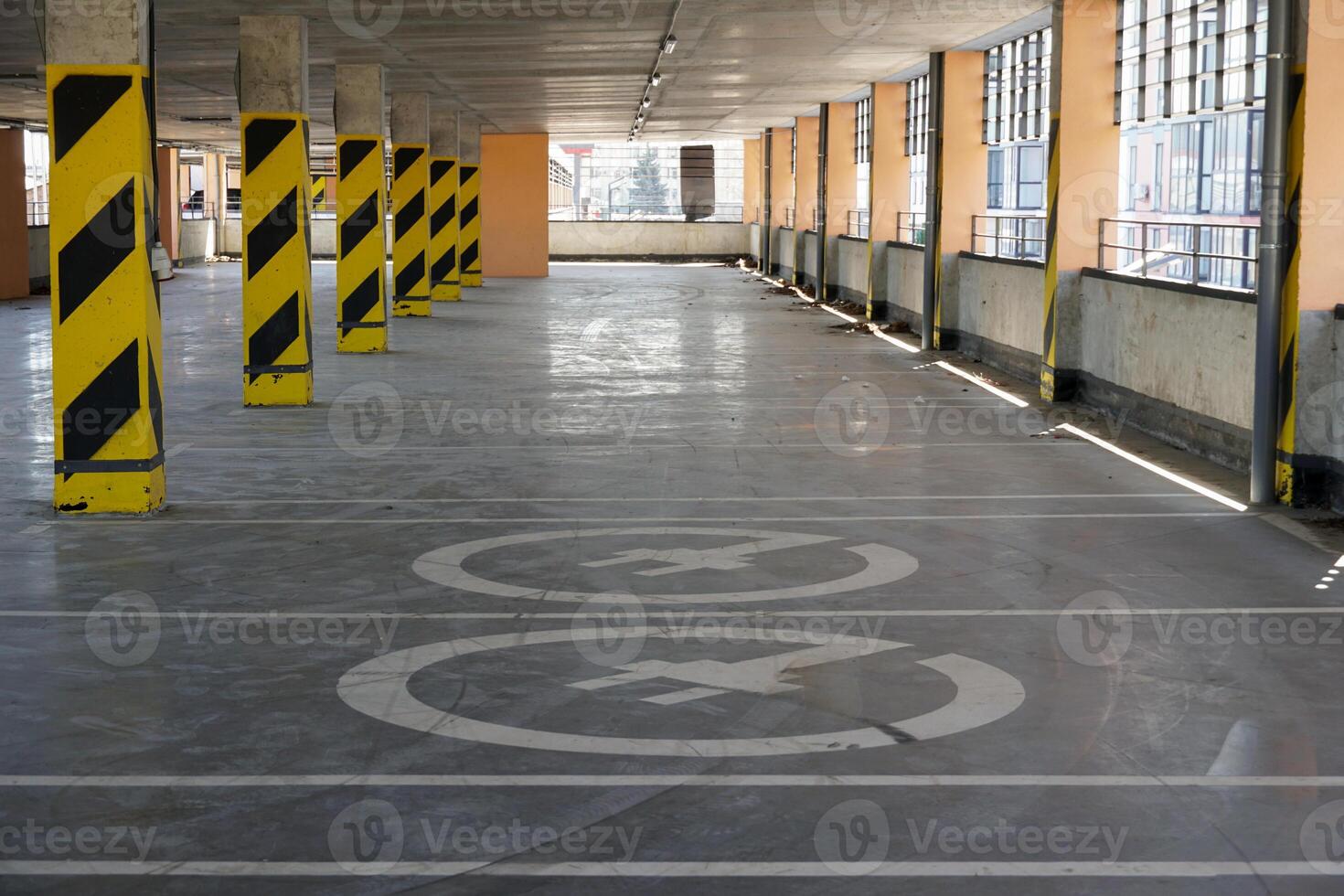 A spacious parking garage features clear directional markings with yellow stripes. Natural light filters in through large windows, creating a bright atmosphere photo