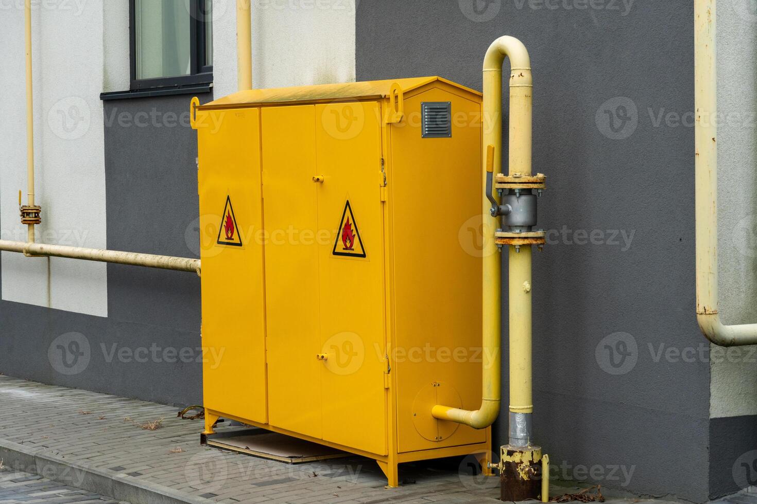 Utility cabinet painted in bright yellow is placed beside a gray wall, featuring warning signs. Pipes run along the structure, indicating safety measures and functionality photo