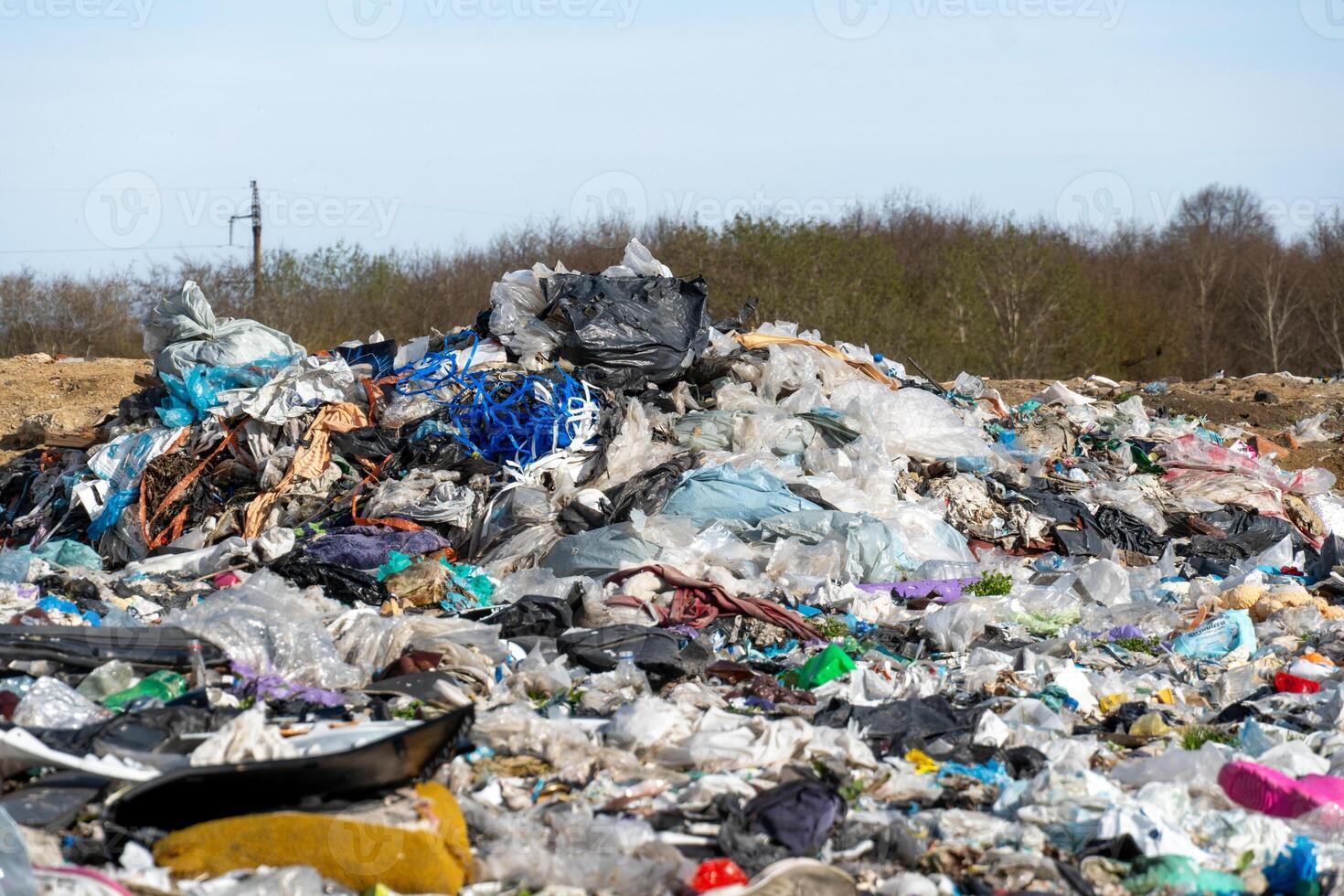 A large pile of various discarded materials, primarily plastic, accumulates in a landfill site under clear skies. Surrounding vegetation indicates an urban area nearby photo
