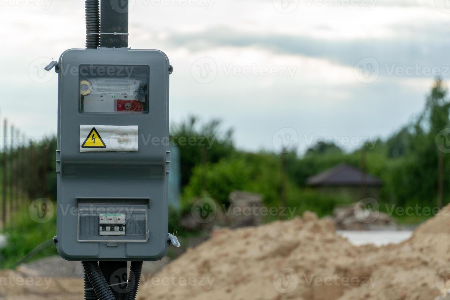 A new electrical meter box is mounted on a pole at a construction site surrounded by sand and greenery under overcast skies photo