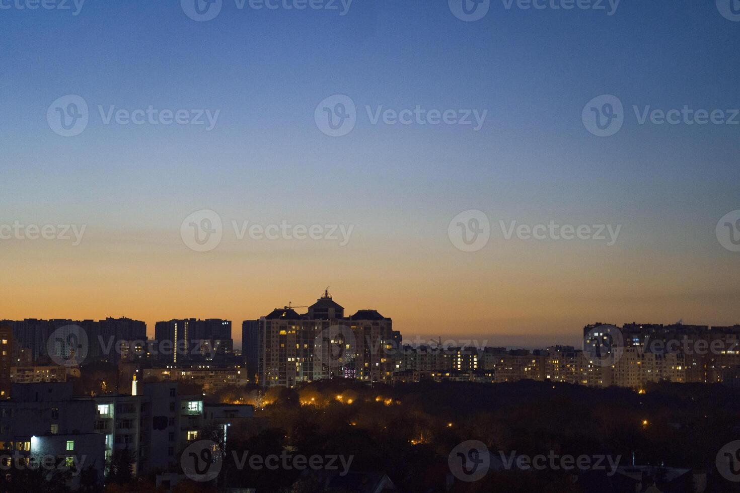 Urban skyline at dusk, showcasing illuminated buildings against a gradient sky transitioning from orange to deep blue, creating a serene evening atmosphere with city lights photo