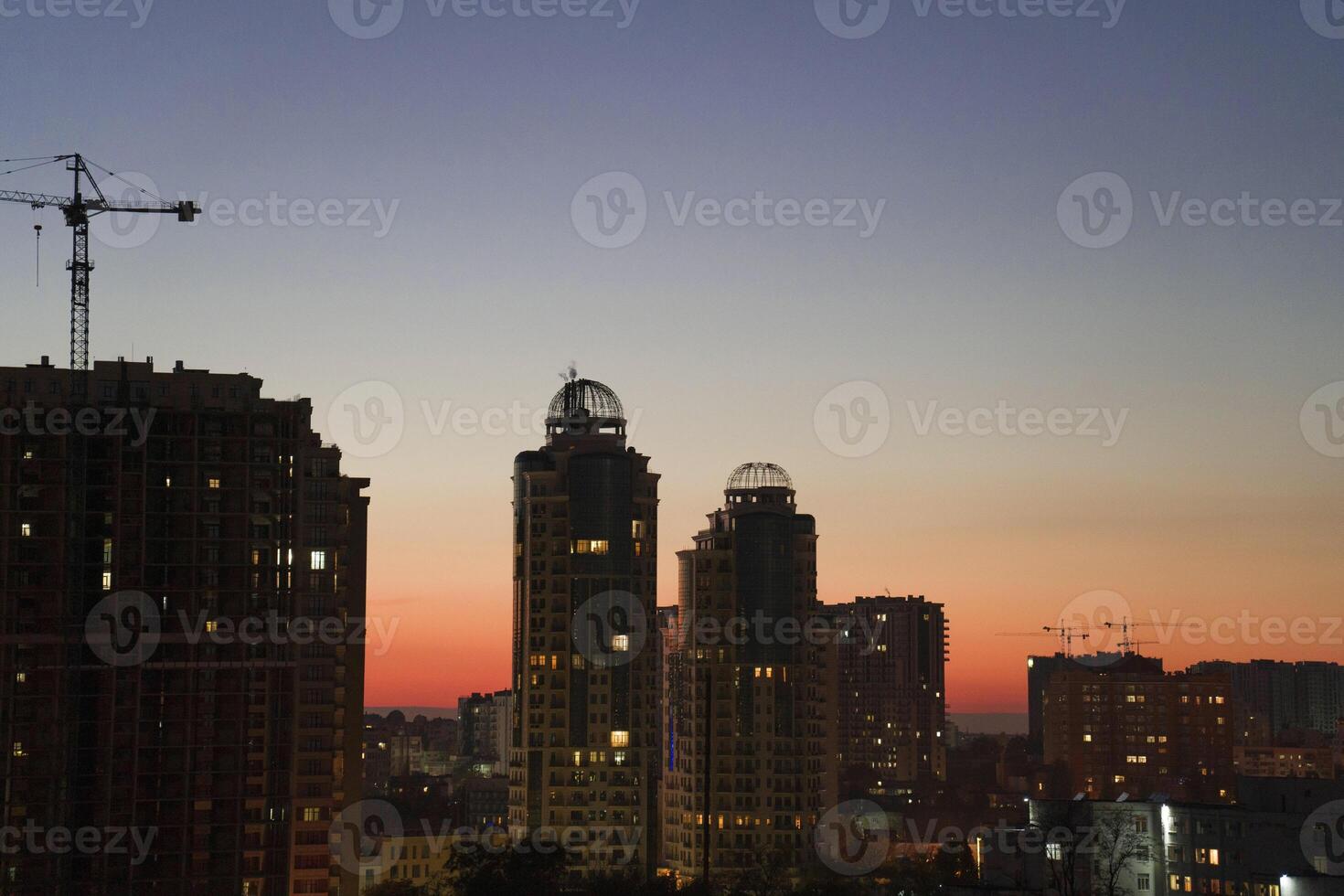 Urban skyline at dusk featuring modern high-rise buildings with illuminated windows, construction crane, and a vibrant sunset sky creating a stunning cityscape atmosphere photo