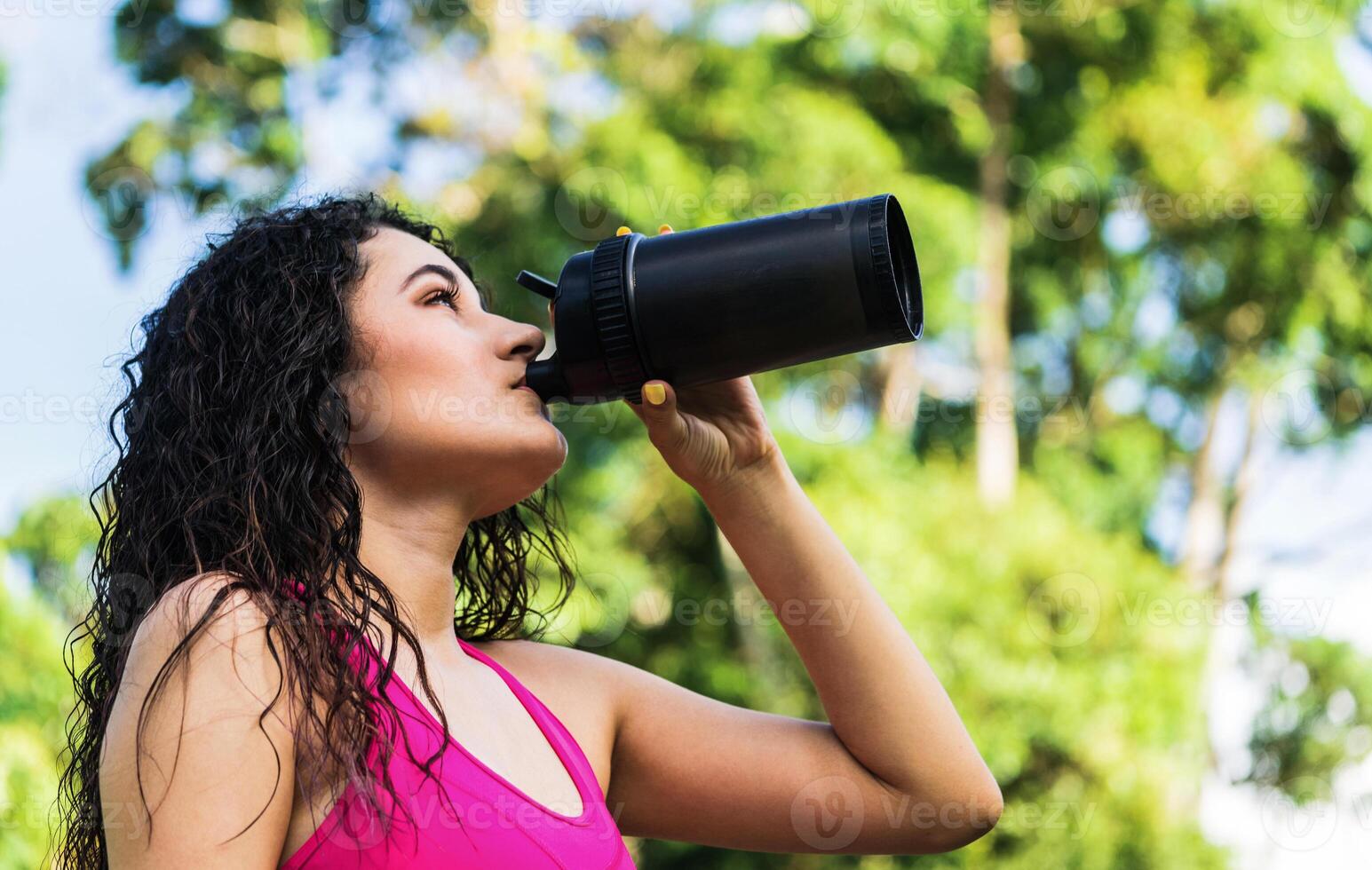 a young latinx woman in sportswear holds a bottle of drink while drinking. young hispanic woman drinking water. photo