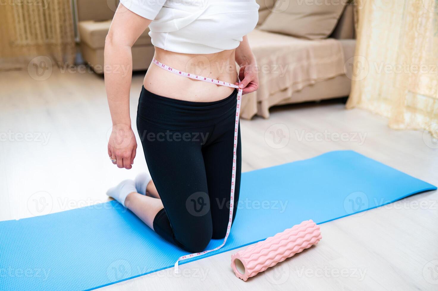 Woman with imperfect waist measuring it with tape while standing on yoga mat, body awareness photo