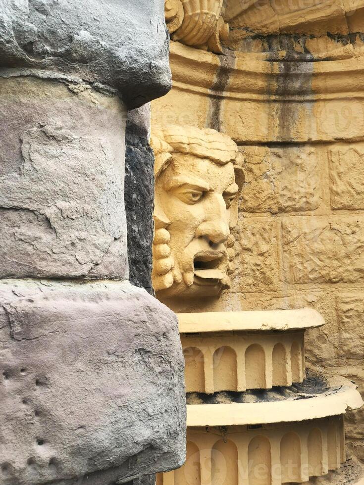 Classical stone sculpture in a historic fountain. The sculpted face, with intricate details, is set against a weathered yellow wall, partially hidden by rough stone blocks. photo