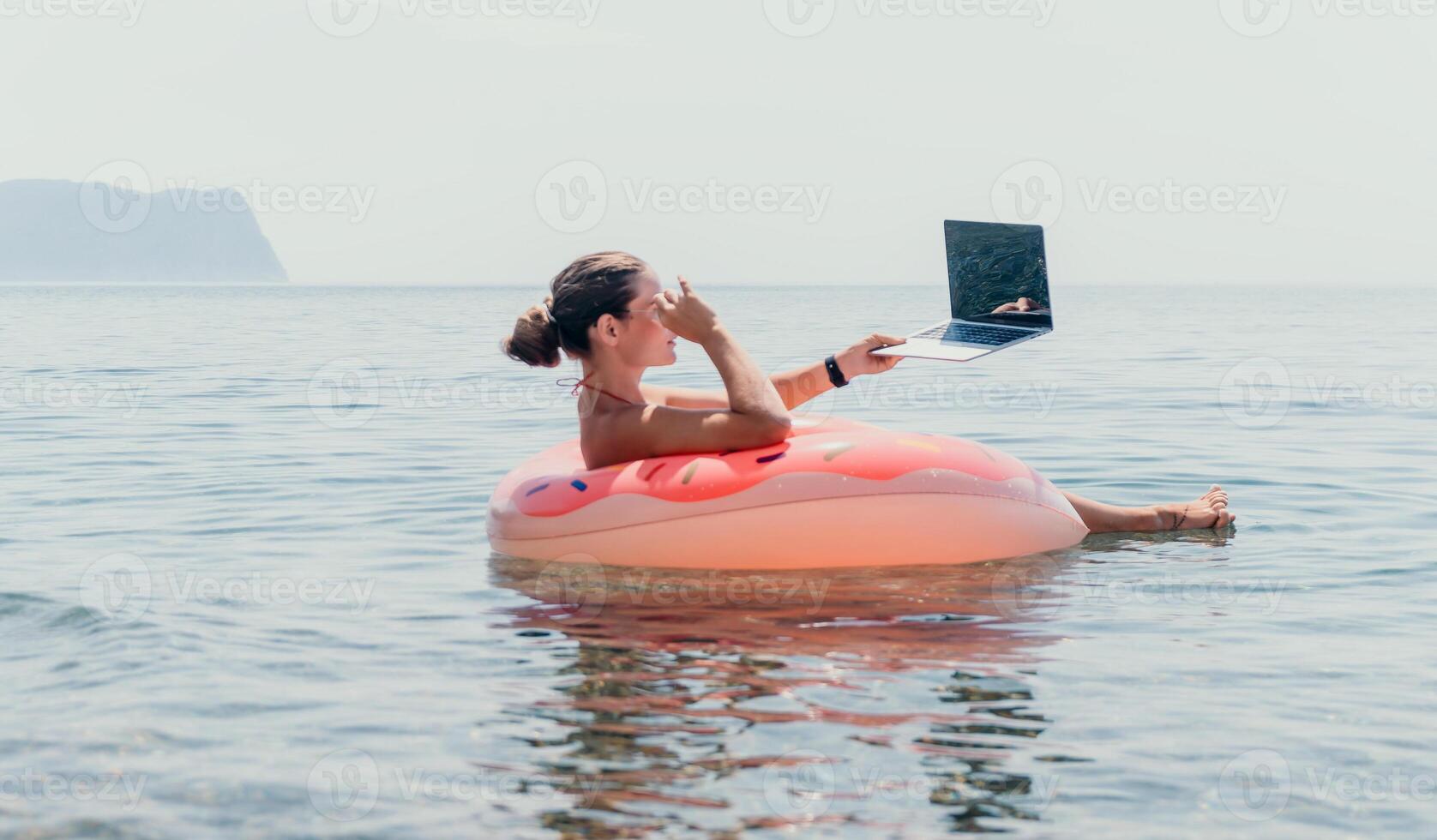 Woman, Beach, Laptop - Working remotely while floating on a pink inflatable ring in the sea. photo