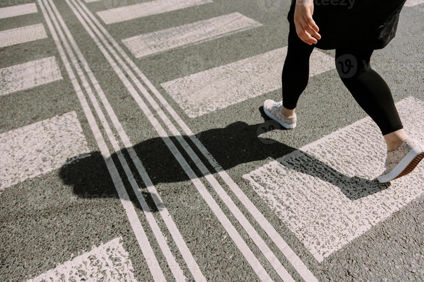 A woman walking briskly across a sunlit crosswalk with a sharp shadow falling on the pavement. Ideal for themes of lifestyle, urban movement, freedom, or motivation. photo