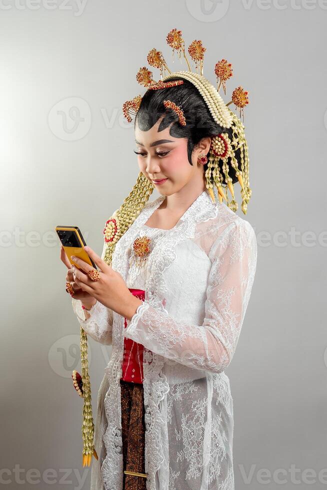 Indonesian bride in Solo Putri attire elegantly interacts with her smartphone. Wearing a golden headdress, jasmine accessories, and a lace kebaya, she smiles while looking focused. photo
