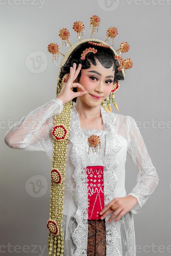 Indonesian bride in Solo Putri attire wearing a lace kebaya, intricate headdress, and jasmine garlands. She smiles gracefully while making an OK hand gesture. photo