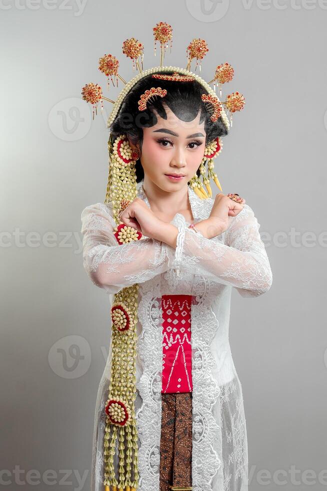 A Indonesian bride in Solo Putri attire, wearing an ornate headdress and lace kebaya. She crosses her arms in an X gesture with a determined expression, exuding strength and elegance photo