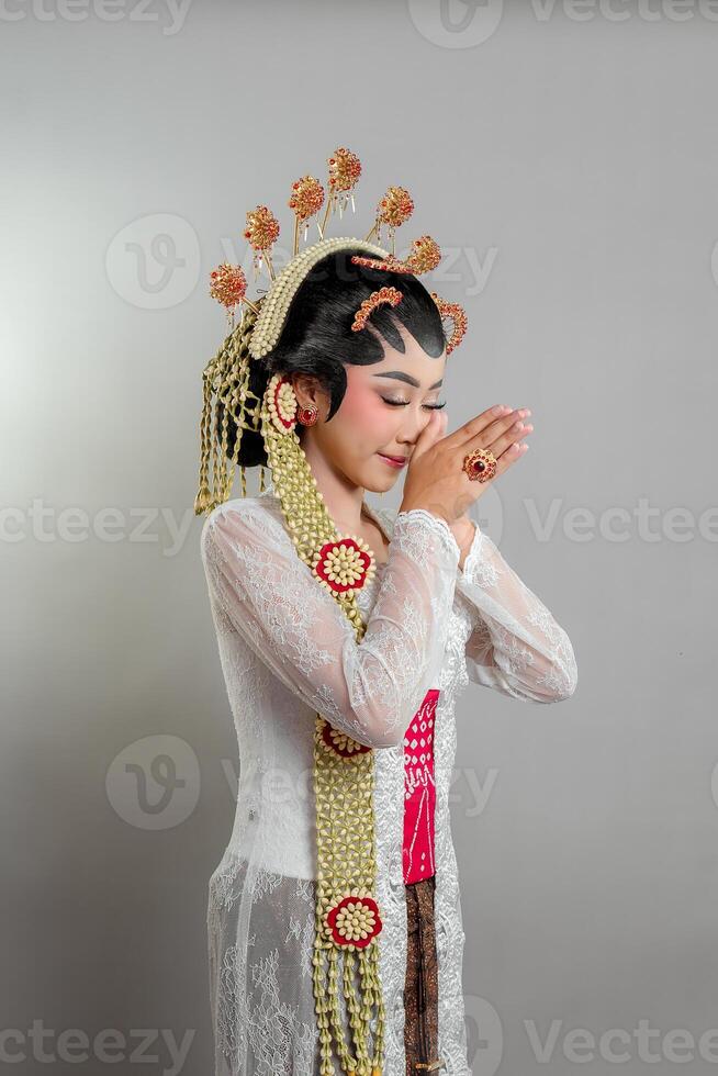 Indonesian bride Solo Putri attire stands gracefully with a soft smile, hands in a respectful greeting. She wears a golden headdress, jasmine garland, and a white lace kebaya photo