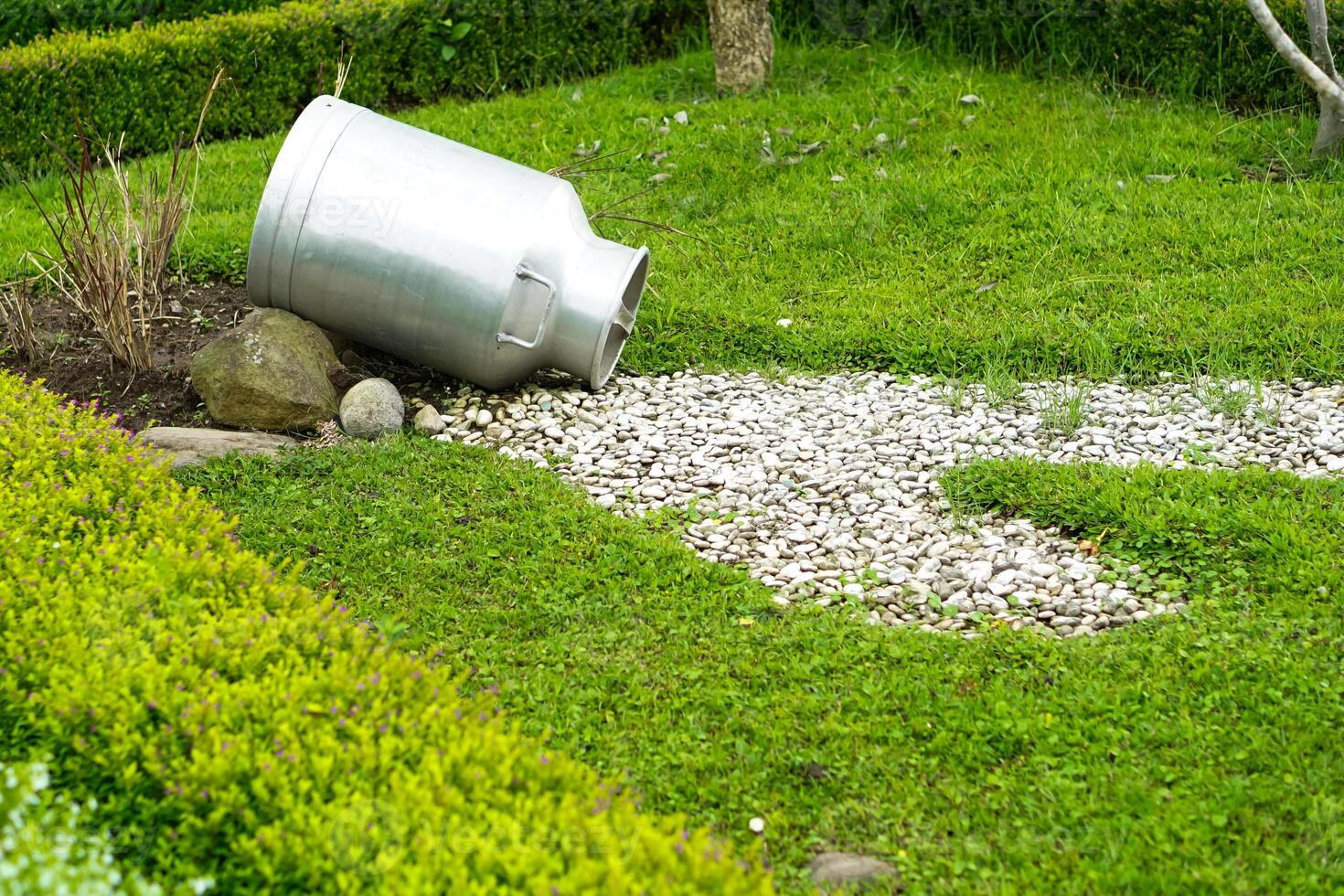 An old metal milk can placed on green grass with white pebbles arranged like a flowing spill, creating a rustic garden decoration that evokes countryside charm and creativity. photo