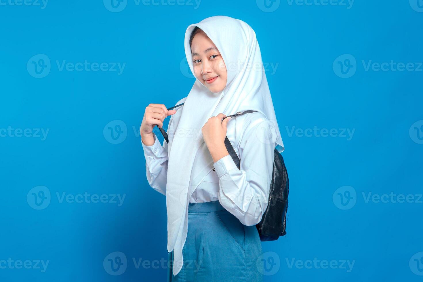 Cheerful Asian teenage girl wearing school uniform and hijab, smiling confidently while holding backpack straps. Positive and friendly facial expression on blue background. photo