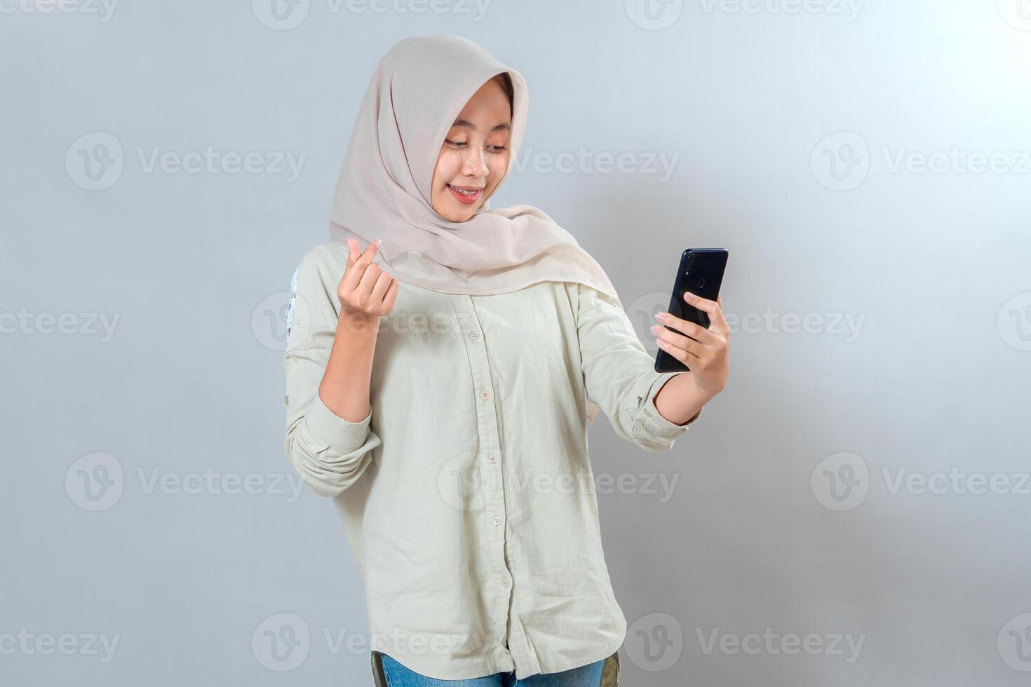 Cheerful young Asian woman in hijab smiling while taking a selfie and making a finger heart gesture. Bright facial expression, confident and friendly vibe, studio shot. photo