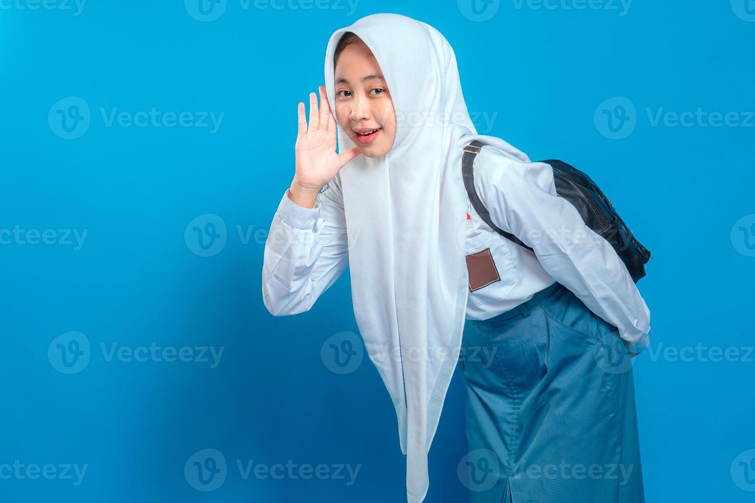 A Cheerful Muslim schoolgirl in uniform and hijab making a listening gesture with playful smile and expressive face. Back to school theme on isolated blue background. photo