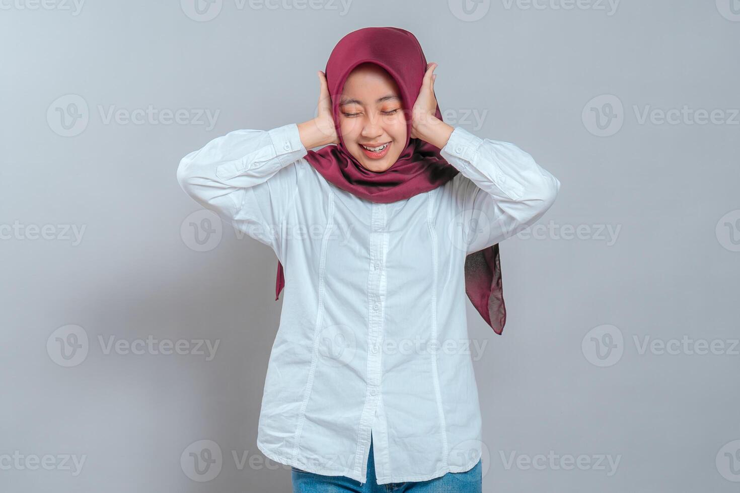 Happy young Muslim woman in hijab smiling with eyes closed, covering her ears playfully. Cheerful facial expression and lighthearted mood perfect for advertising or lifestyle content. photo