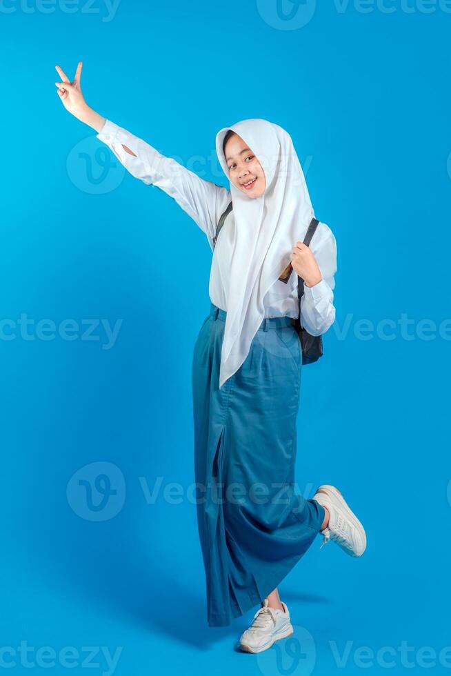 A Asian Muslim teenage girl smiling cheerfully with a happy and confident expression, showing a peace sign gesture while standing on one leg, back to school education concept. photo