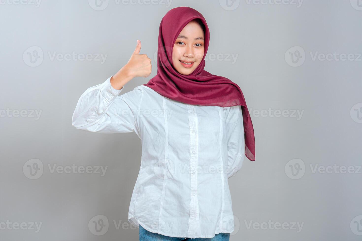 A Confident Muslim woman wearing a maroon hijab smiling warmly while giving a thumbs-up gesture. Her bright expression conveys positivity, approval, and encouragement. photo