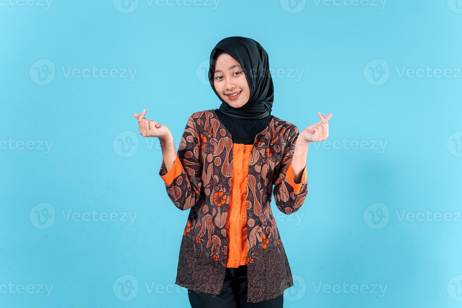 Smiling young Asian woman wearing a hijab and traditional batik outfit makes a finger heart gesture, expressing joy and friendliness in a studio portrait. photo