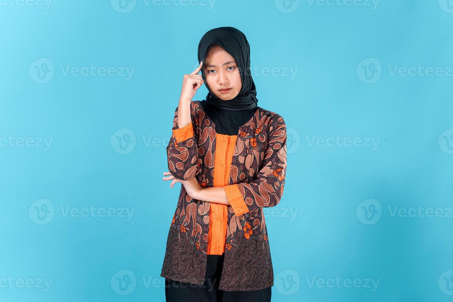 A Young Asian woman wearing hijab and batik outfit looks thoughtful and serious, with one hand on her chin and the other pointing to her temple, expressing deep thinking or concern. photo