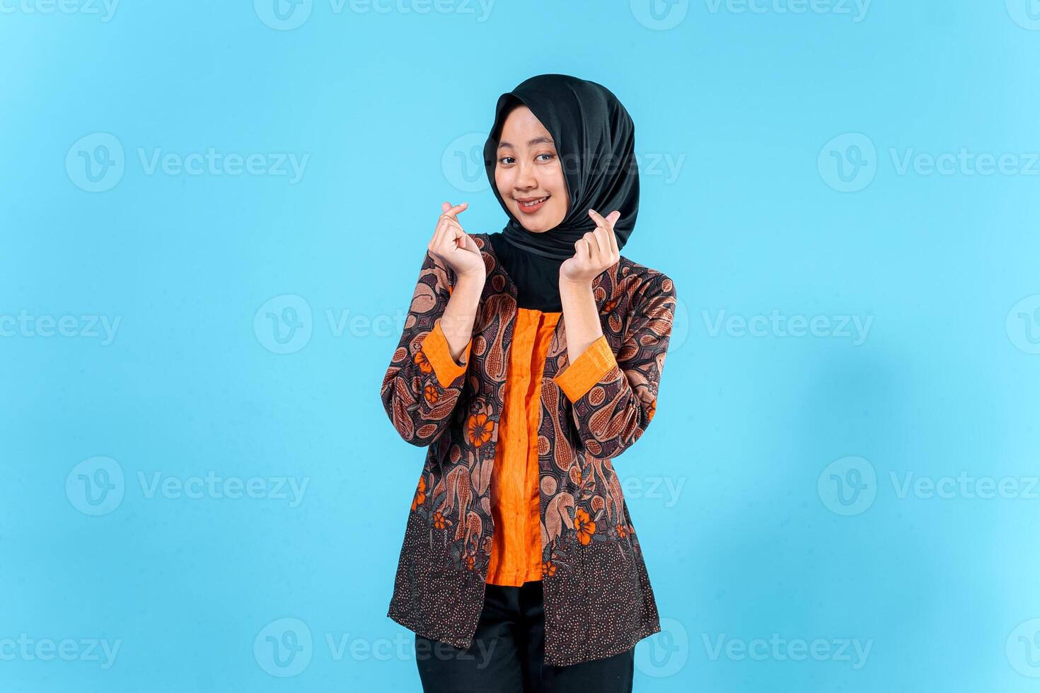A Smiling Asian woman wearing a hijab and traditional batik outfit makes a finger heart gesture, expressing joy and friendliness in a studio portrait. photo