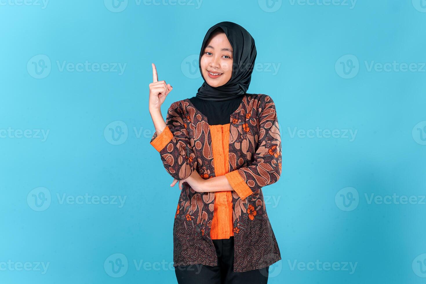 Confident young Asian woman wearing hijab and batik outfit smiling brightly while pointing upward with one finger, expressing excitement, inspiration, or having a great idea. photo