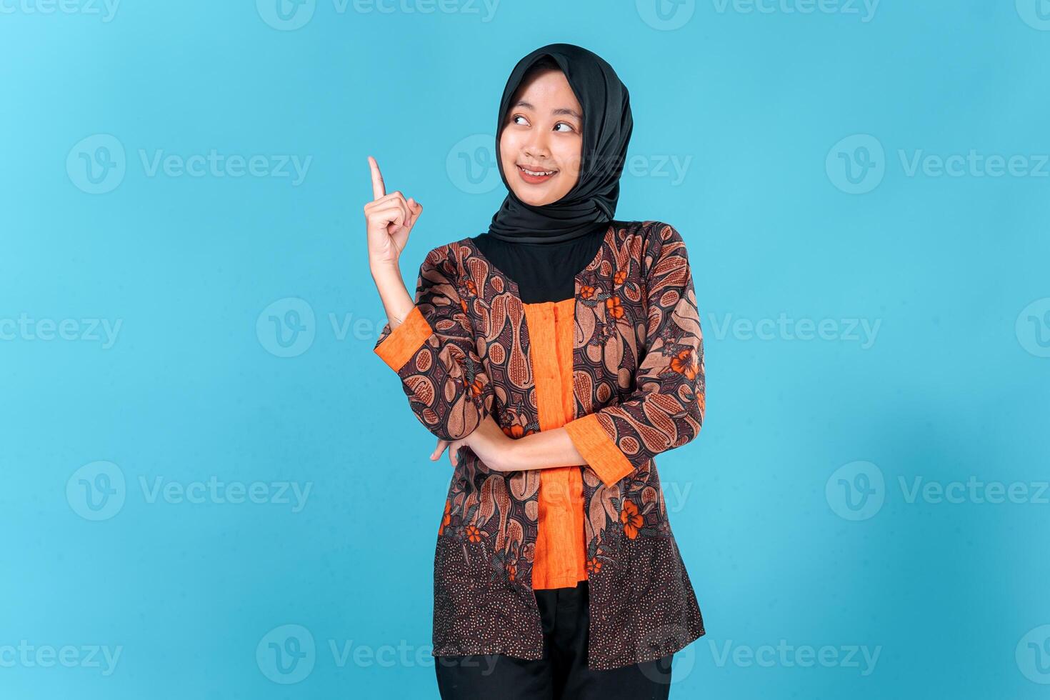 A Confident young Asian woman wearing hijab and batik outfit smiling brightly while pointing upward with one finger, expressing excitement, inspiration, or having a great idea. photo