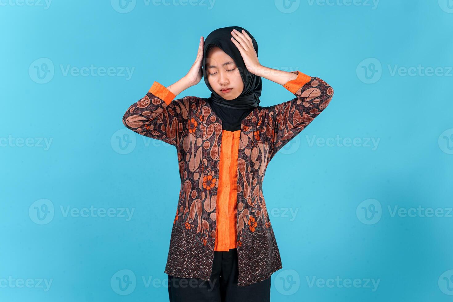 A Young Asian woman wearing hijab and traditional batik clothes looks stressed and frustrated, holding her head with eyes closed, expressing headache or mental fatigue. photo