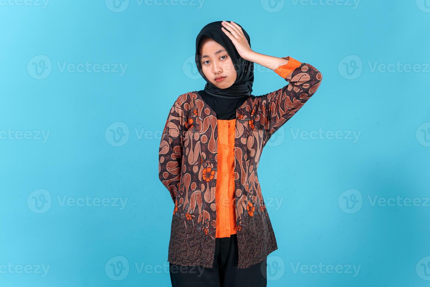 Young Asian woman wearing hijab and traditional batik clothes looks stressed and frustrated, holding her head with eyes closed, expressing headache or mental fatigue. photo
