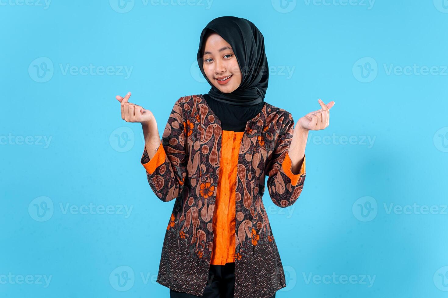 A young Asian woman wearing a hijab and traditional batik outfit makes a finger heart gesture, expressing joy and friendliness in a studio portrait photo