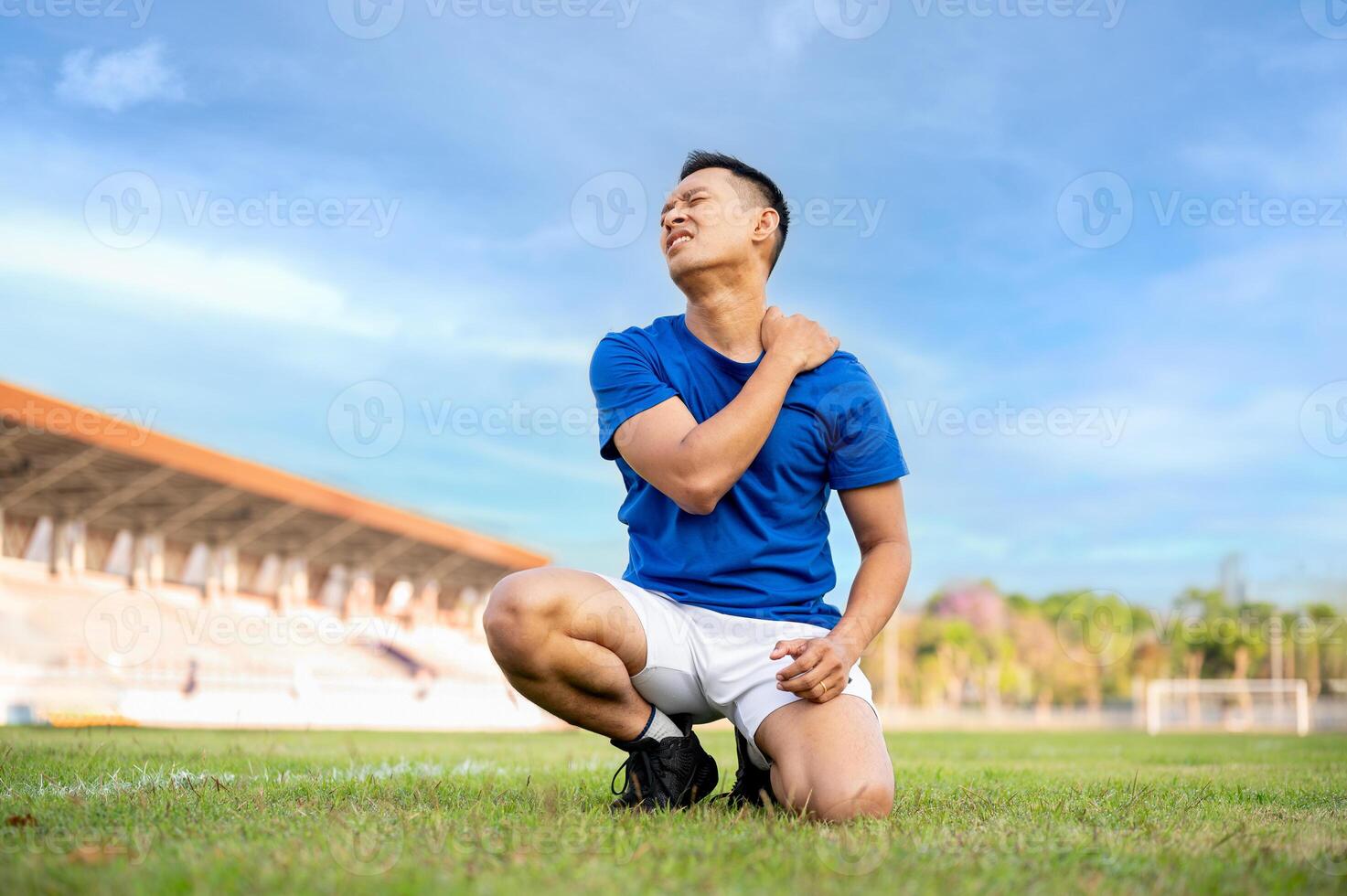 Asian man in blue shirt is kneeling on grass and touching or massaging his shoulder. Pain or injury. photo