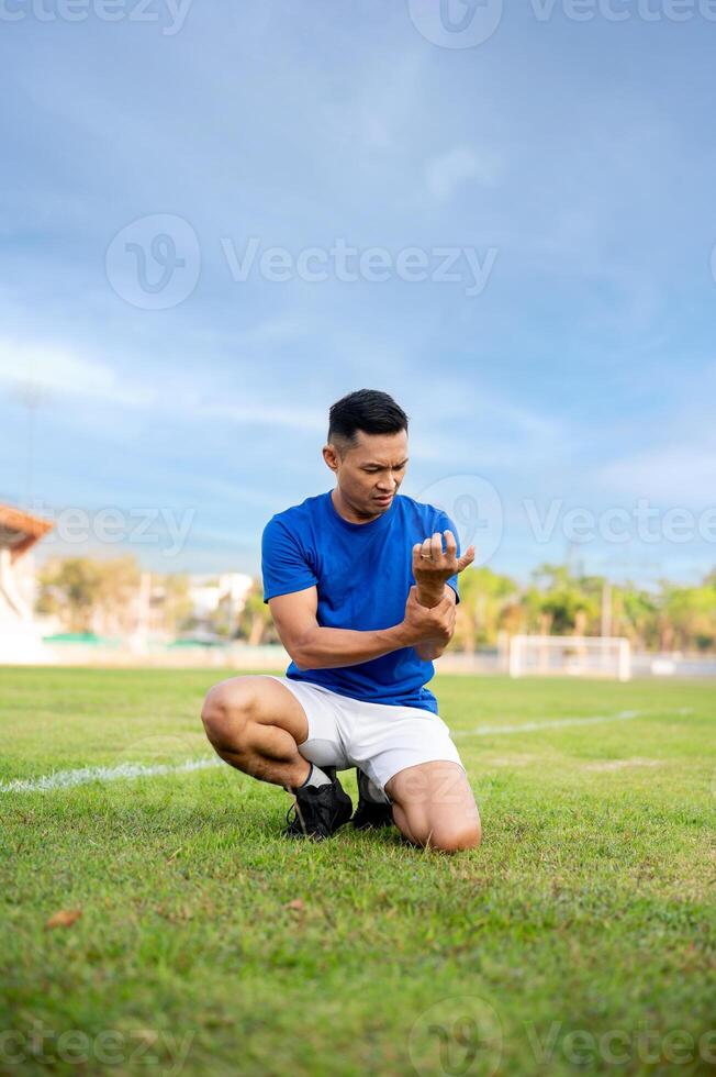 Asian man in blue shirt holding and looking at his arm while kneeling on grass. Arm pain or injury. photo