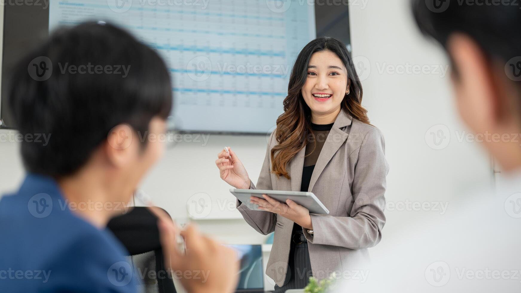 Smiling businesswoman or office worker holding digital tablet while speaking to coworkers in meeting photo