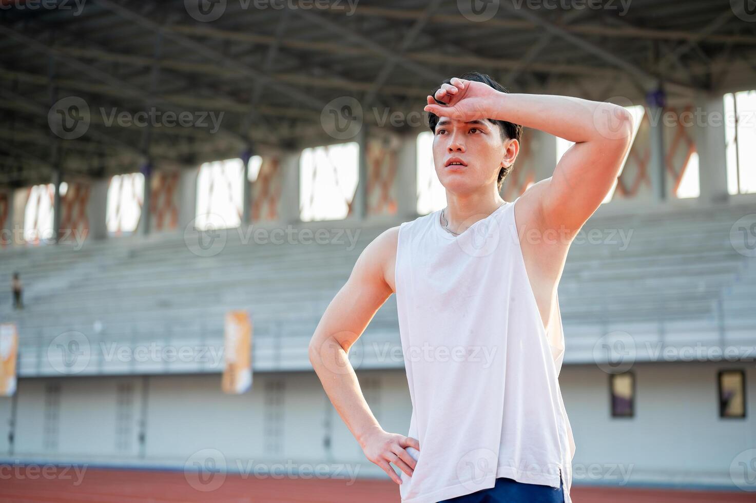 Asian man in white tank top standing with hand on waist and wiping sweat after running on racetrack. photo