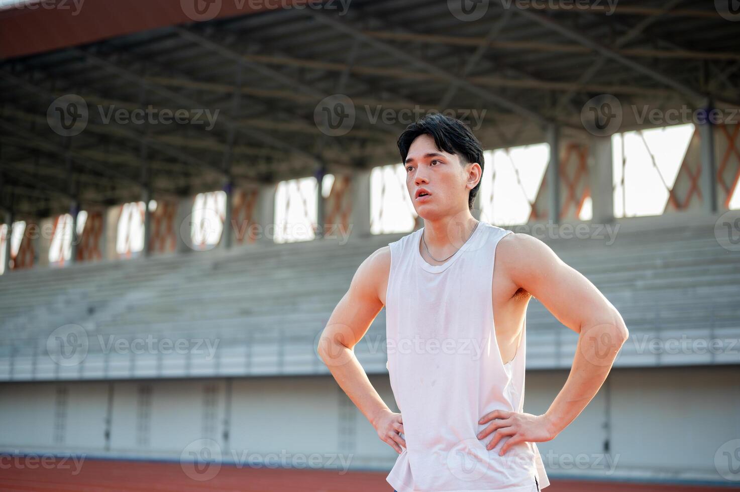 Asian man in white tank top is standing with hands on waist taking a rest after running on racetrack photo