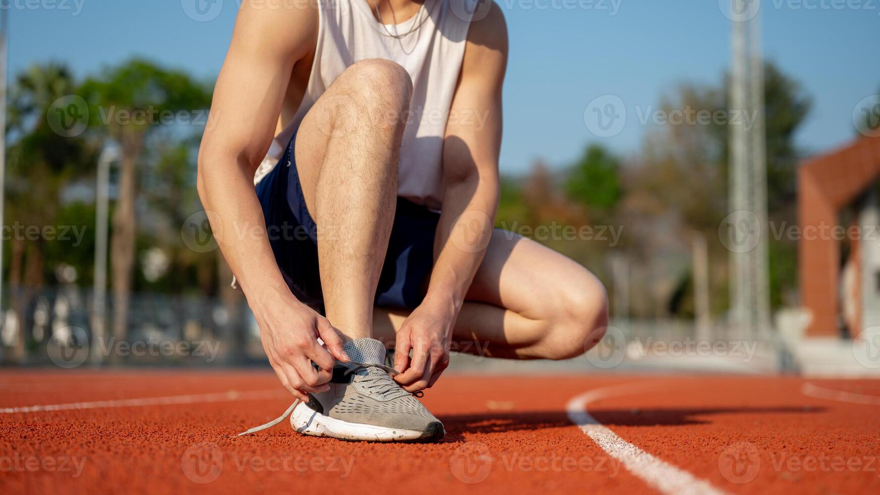 Close up of a man in white tank top crouching while tying his sneaker shoelace on the racetrack. photo