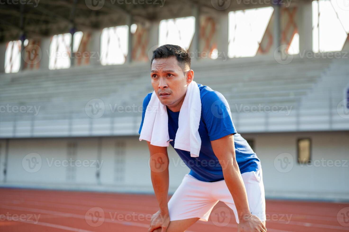 Asian man in blue t shirt standing with hands on his knee, taking a rest after running on racetrack. photo