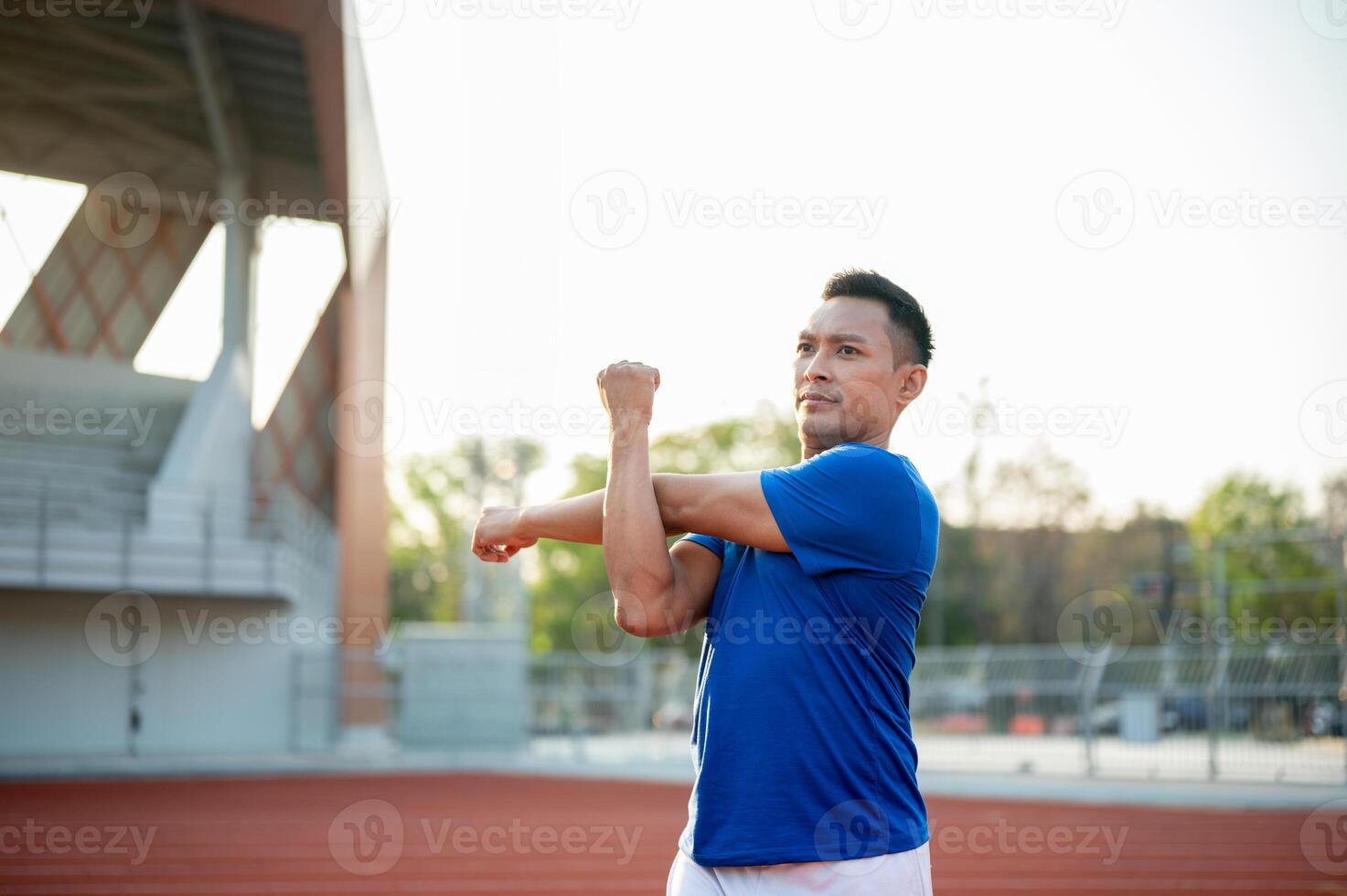An asian man in blue shirt stretching his arm and shoulder muscles on racetrack. Outdoor exercise. photo