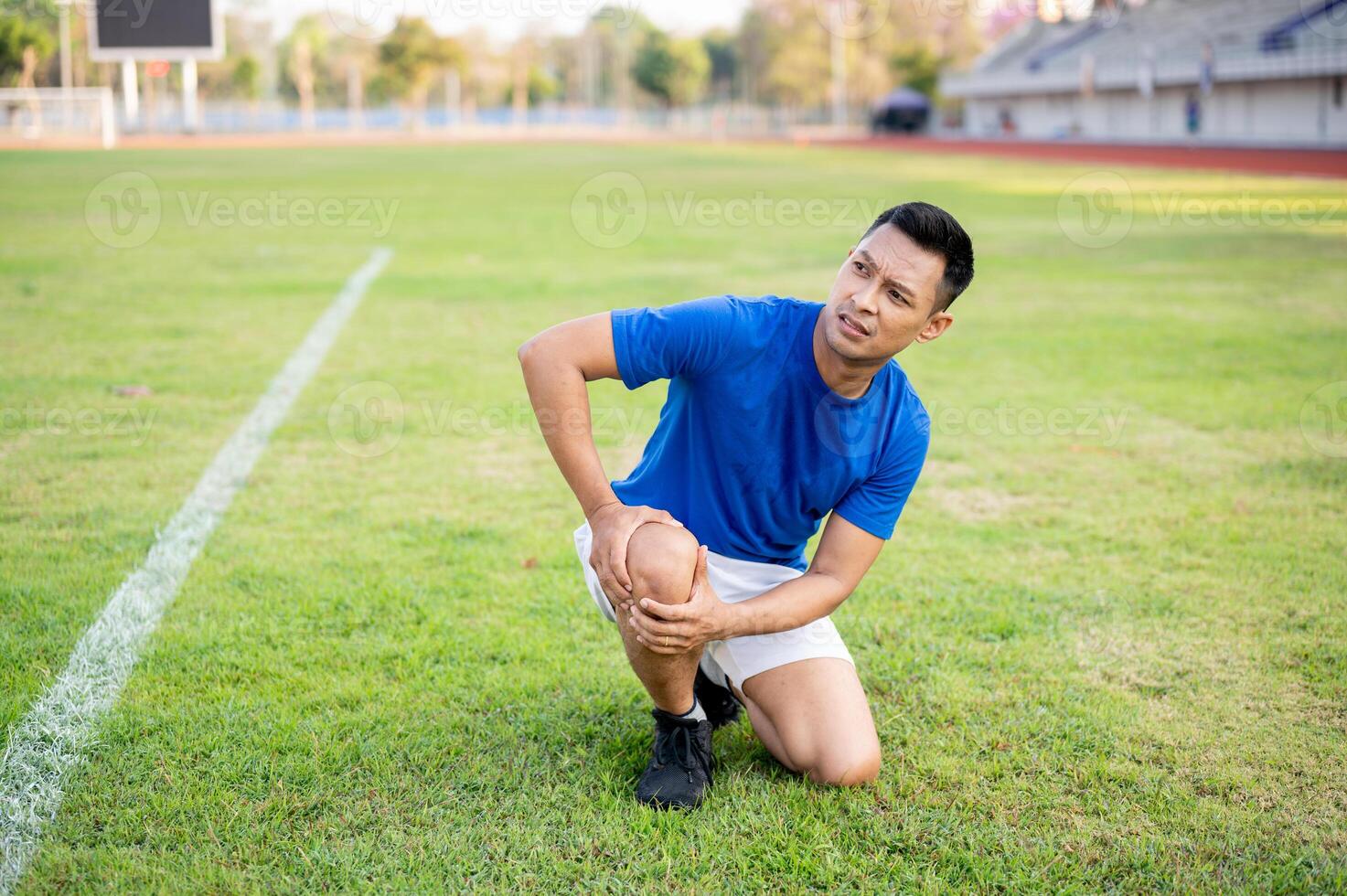 Asian man in blue shirt kneeling on grass and holding his knee while looking up. Leg pain or injury. photo