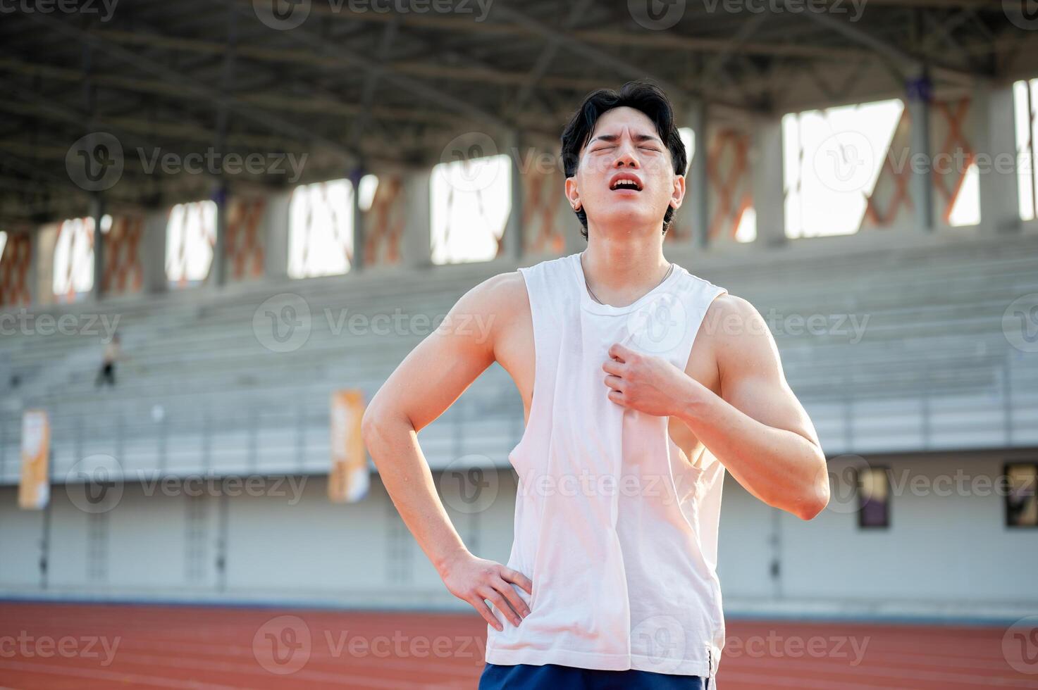 Asian man is standing with one hand on waist and other hand grabbing a white tank top. Hot or tired. photo