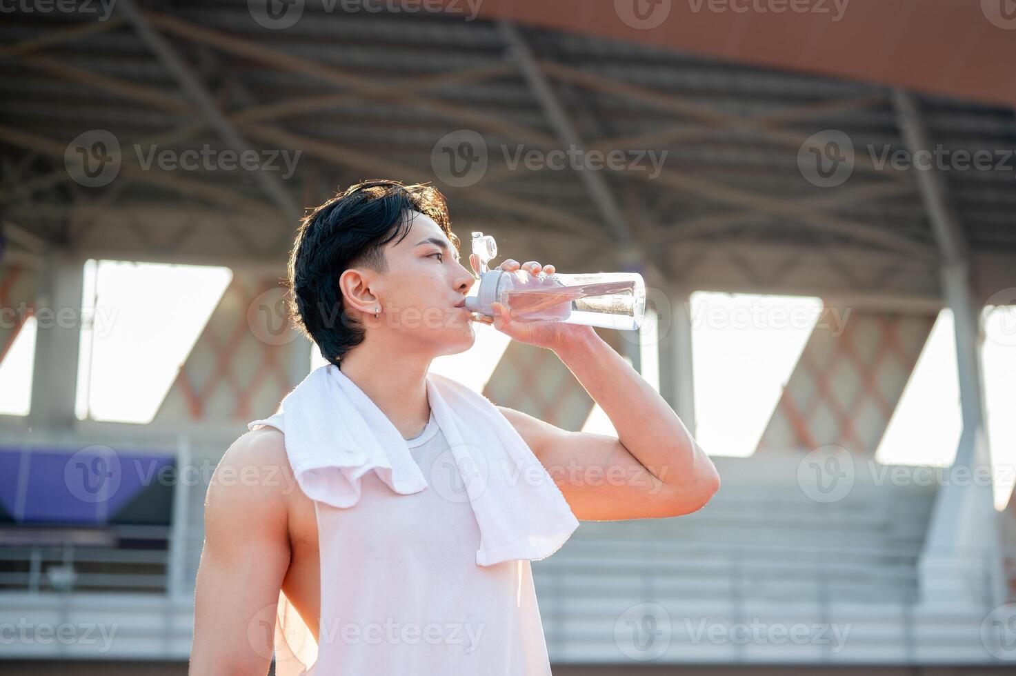 Asian man in white tank top standing and drinking from a water bottle, taking a rest after running. photo